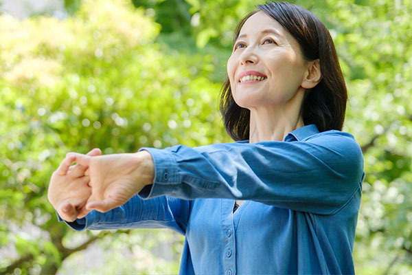 屋外の新緑の中で笑顔で伸びをする青いシャツの女性